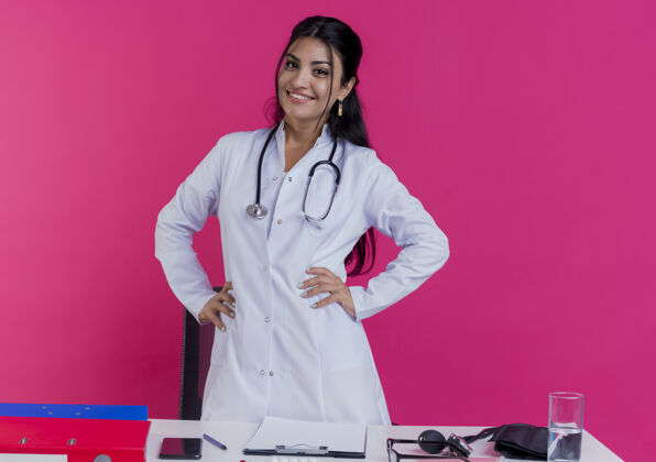 听诊器面带微笑的年轻女医生穿着医用长袍 手持听诊器站在桌子后面 手放在腰上 隔离在粉红色的墙上医生看年轻