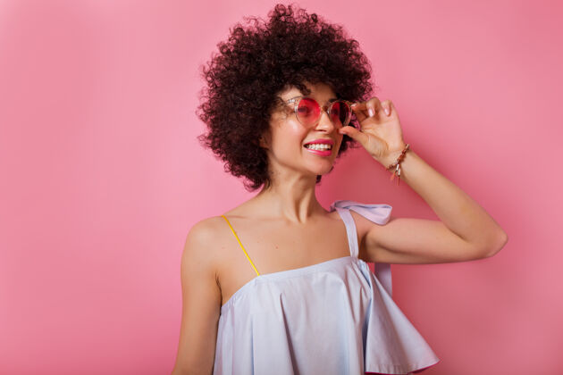 大笑兴奋的时尚女孩 卷发 穿着浪漫的夏季衬衫 戴着圆粉色眼镜 微笑着看着粉色的墙壁眼睛健康治疗