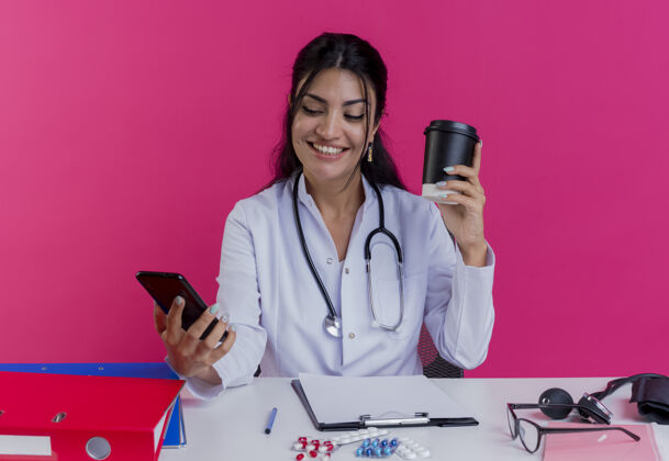 抱着面带微笑的年轻女医生穿着医用长袍和听诊器坐在办公桌旁拿着医疗工具拿着咖啡杯和手机看着隔离在粉红色墙上的手机微笑医疗咖啡