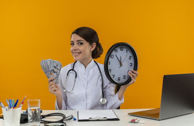 医生面带微笑的年轻女医生穿着医用长袍 听诊器坐在办公桌旁 手里拿着医疗工具和笔记本电脑 黄色的墙上孤立着时钟和钞票时钟办公桌坐着