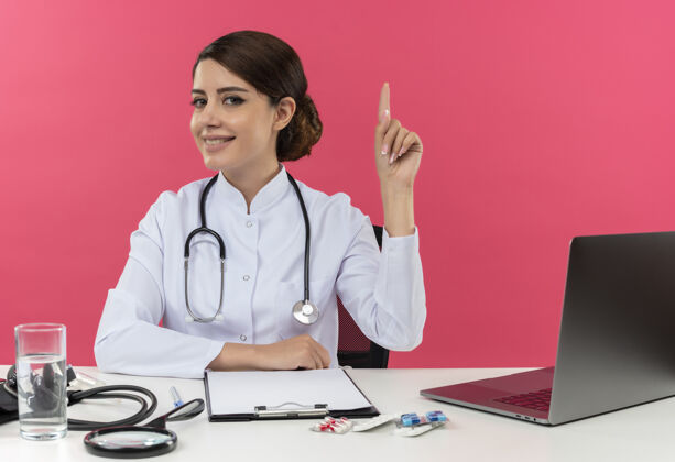 长袍面带微笑的年轻女医生穿着医用长袍和听诊器坐在办公桌旁 手里拿着医疗工具和笔记本电脑 把手放在办公桌上 孤立地指着粉红色的墙上微笑医疗手