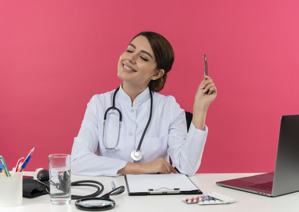 办公桌面带微笑的年轻女医生穿着医用长袍和听诊器坐在办公桌旁 手里拿着医疗工具和笔记本电脑 手里拿着信用卡 眼睛紧闭 隔离在粉红色的墙上医疗听诊器女