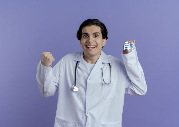 长袍快乐的年轻男医生穿着医用长袍和听诊器 在紫色的墙上显示一包胶囊在做“是”的手势胶囊听诊器手势