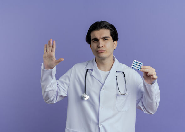 男性皱眉的年轻男医生穿着医用长袍和听诊器 显示一包胶囊正在做停止手势 隔离在紫色的墙上 留有复印空间年轻看胶囊