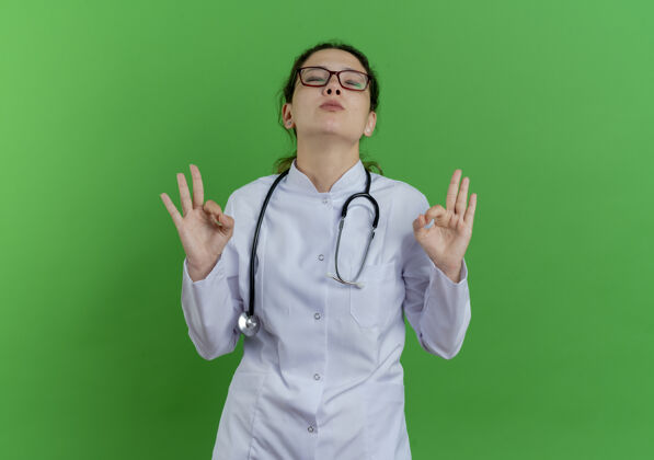 和平和平的年轻女医生穿着医用长袍 戴着听诊器 戴着眼镜 闭着眼睛做着“ok”的手势 隔离在绿色的墙上 留着复印空间年轻医疗封闭
