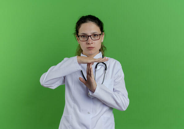 女严格的年轻女医生穿着医用长袍 戴着听诊器和眼镜 在绿色的墙壁上做着暂停动作 并留有复印空间听诊器年轻手势
