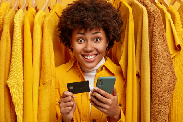 技术正面混血购物狂站在衣架旁 用信用卡和智能手机购物时尚衣服销售