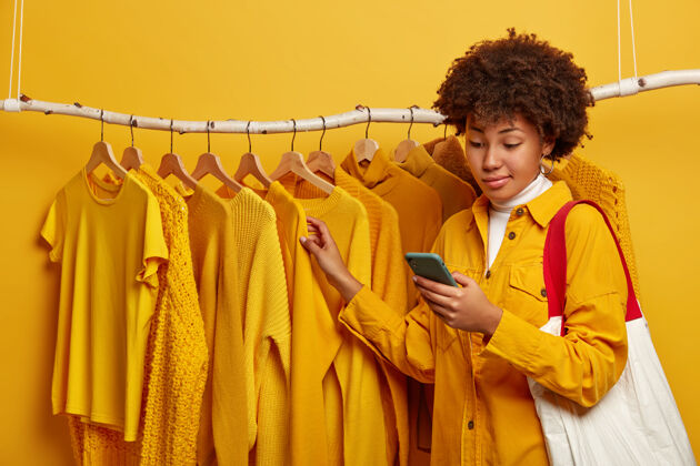 电子不知情的黑皮肤非洲发型女人 站在黄色衣架旁 肩上扛着购物袋 挑选新装行装女人室内