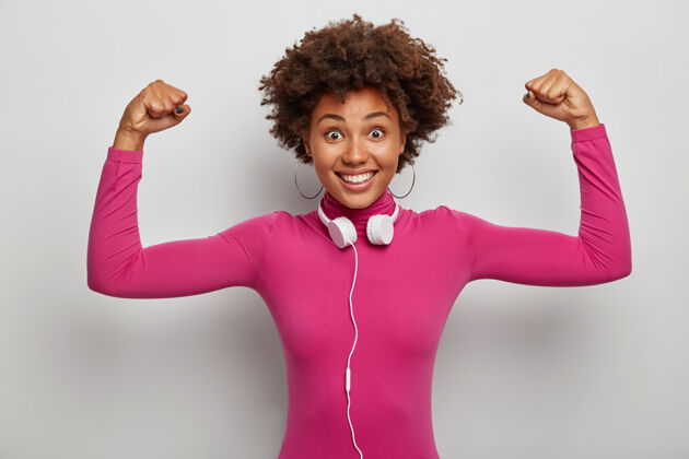 休闲精力充沛的美国黑人女士举起双臂展示肌肉和力量 笑容灿烂 脖子上戴着立体声耳机微笑力量人