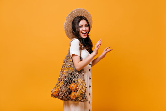 包卷发的时髦女人拿着带水果的环保袋摆姿势戴草帽的女孩在橙色背景上微笑微笑女孩化妆品