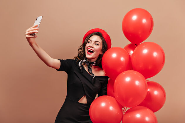 优雅身穿黑色连衣裙 头戴红色贝雷帽的可爱女子手持智能手机 自拍 并与气球合影女士米色背景漂亮