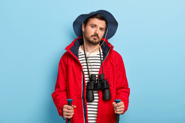 白天严肃的年轻男性徒步旅行者在树林里旅行 使用登山杖 享受旅游生活方式 脖子上戴着望远镜 戴着休闲帽和红色夹克 隔离在蓝色的墙上旅行站立徒步旅行