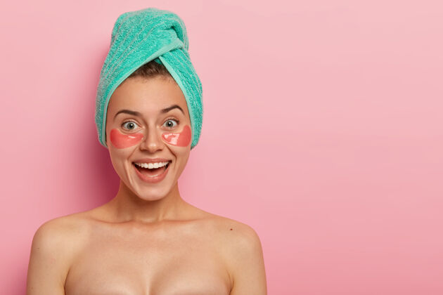 室内积极的女人戴着胶原蛋白贴片在眼睛下面 有美容治疗 赤身裸体站在室内 有广泛的微笑 迷人的外观皮肤护理肖像皮肤科