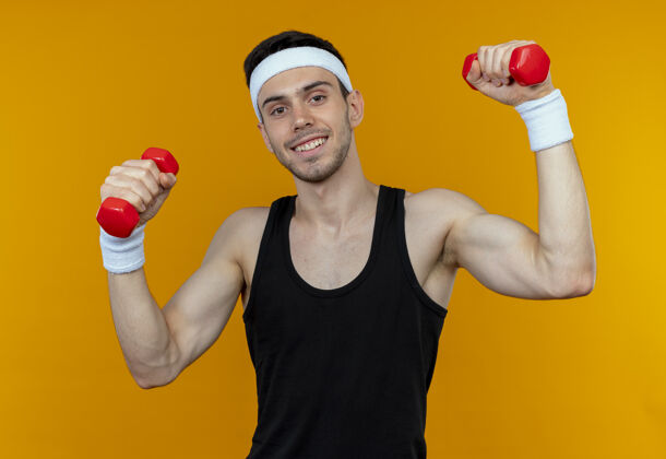 头带戴着头巾的年轻运动型男子 带着哑铃在橘子上微笑着锻炼身体哑铃微笑年轻