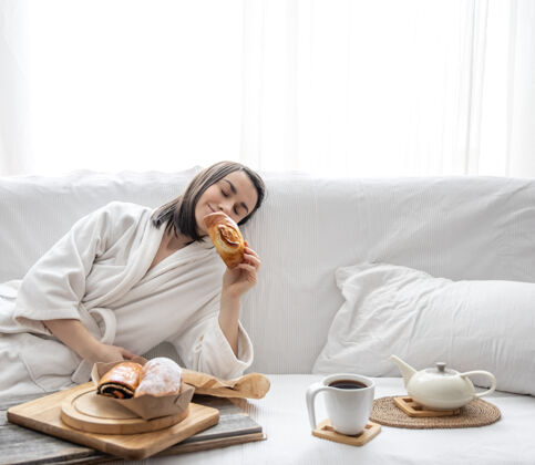 早餐一个可爱的年轻女子穿着长袍坐在沙发上 手里拿着一个发髻早午餐早晨面包