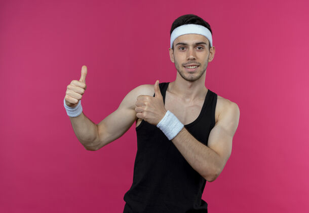微笑戴着头巾的年轻的运动型男人微笑着快乐而积极地向粉色竖起大拇指拇指积极站