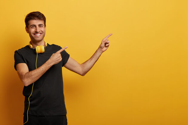 耳机帅气开朗的男人指着复制空间 穿着黑色衣服 戴着耳机 牙牙学语地笑着 展示着广告 隔离在黄色的背景上娱乐听力设备