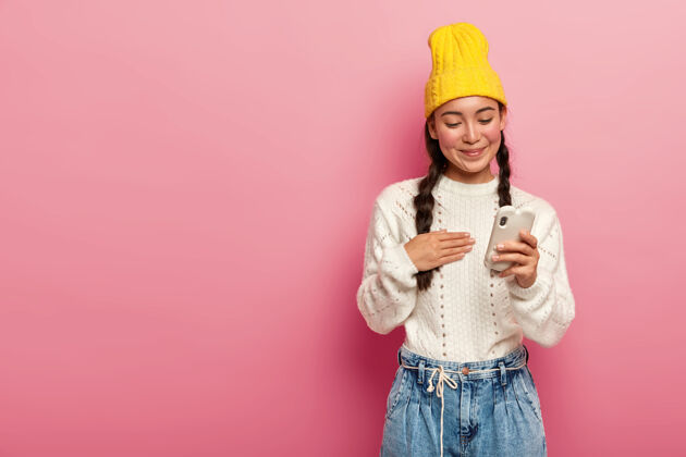 毛衣高兴的深色皮肤的韩国女人用手机在粉红色的背景设备情感新闻