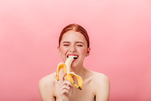 年轻快乐的姜女孩在吃香蕉快乐的女人在粉红色背景下享受热带水果的正面视图新鲜女孩一个人