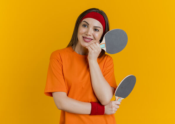 拍面带微笑的年轻运动女性戴着头带和腕带拿着乒乓球拍把手放在下巴上看着侧面放乒乓球运动