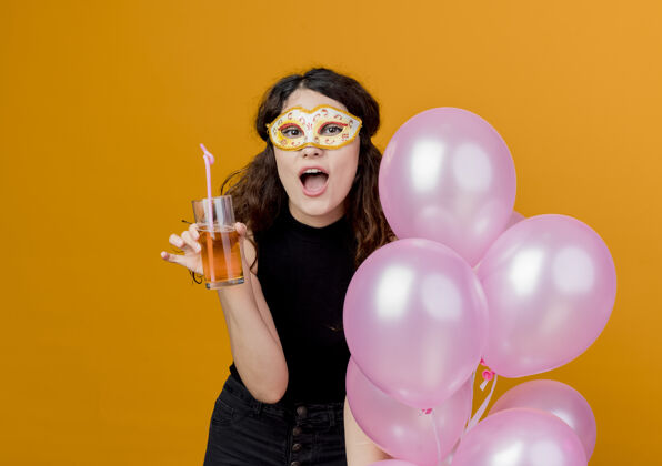 头发年轻漂亮的女士 卷发 手持一束气球和鸡尾酒 戴着派对面具 快乐快乐的生日派对理念 橙色面具空气聚会