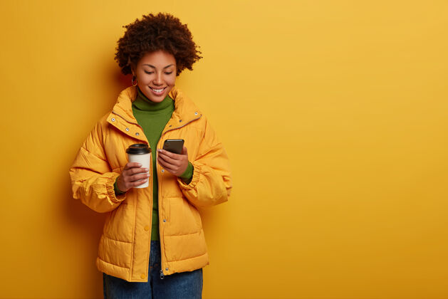 检查穿着黄色外套的有魅力的积极女性 乐于阅读帖子下的好评论 手持现代手机 喝外卖咖啡非洲通知站立
