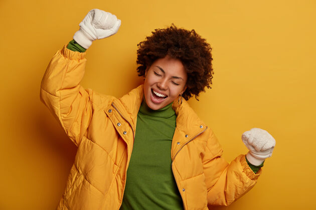 室内喜出望外的年轻美国黑人女子积极地跳舞 穿着暖和的冬衣和手套 有着积极的表情 站在黄色的背景下女人积极快乐