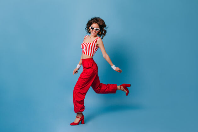 短上衣穿着80年代服装的时髦女人在蓝色的墙上奔跑时尚女士魅力