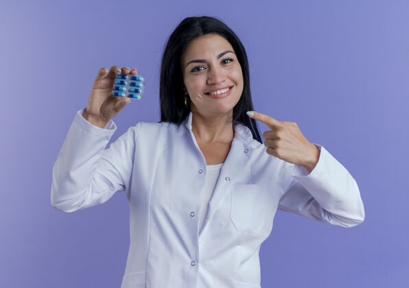 年轻面带微笑的年轻女医生穿着医用长袍 手持并指着一包医用胶囊望着紫色制服健康