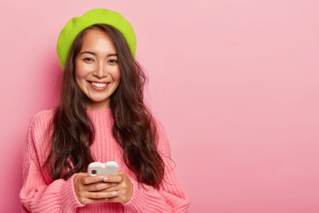 外观微笑的黑发女人 留着长发 戴着亮绿色贝雷帽和超大套头衫 手持现代手机 连接着无线互联网头发时尚人