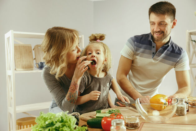 休闲可爱的小女孩和她美丽的父母正在切蔬菜 一边微笑着做沙拉一起积极女人