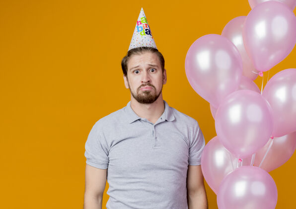 悲伤戴节日帽的年轻人举着气球站在橙色的墙上庆祝生日年轻看帽子