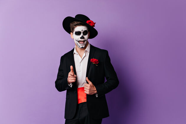 角色穿着僵尸服装的自信男人在紫色背景上摆姿势好看的死人在享受万圣节派对情感骷髅西班牙