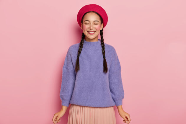 女人满脸欢喜的年轻女子 闭上眼睛 戴着红色贝雷帽 穿着暖和的紫色毛衣和编织的裙子 从接受赞美中得到乐趣 在粉色的墙上摆姿势位置满足室内