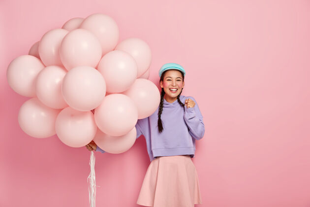 美女快乐的亚洲女孩高兴地握紧拳头 迫不及待地等待着特殊的时刻 在她的生日得到朋友们的祝贺 带着一大串气球 穿着漂亮的衣服模特编织女人