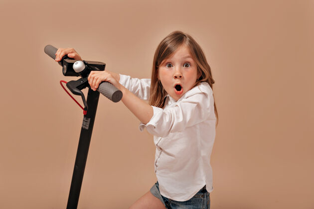 电动滑板车有趣可爱的小女孩穿着白色衬衫与电动滑板车摆出惊喜的情绪情绪小女孩未成年人