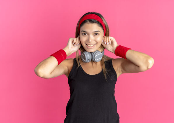 耳机身穿黑色运动服 头戴红色耳机的年轻健身女孩站在粉红色的墙上 开心地微笑着健身女孩年轻