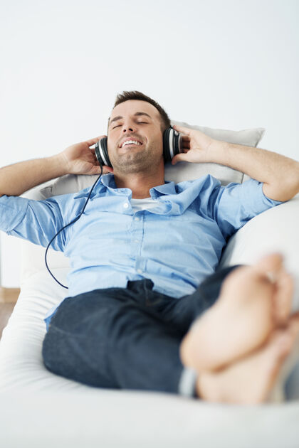 成熟的男人快乐的男人戴着耳机躺在沙发上家居内饰蓝色闭着眼睛