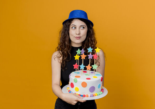 女人一位年轻漂亮的卷发女士 戴着节日礼帽 手里拿着生日蛋糕 面带微笑地站在橙色的墙上脸微笑卷发