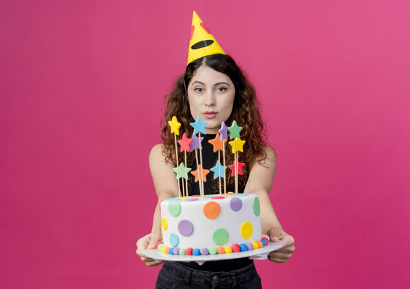 抱着年轻漂亮的女人 卷发 戴着节日帽 手里拿着生日蛋糕 站在粉色的墙上 快乐而积极的生日派对理念帽子女人年轻