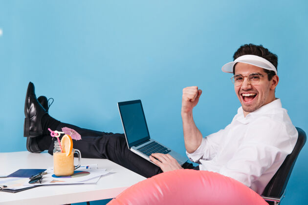 休息一个戴着帽子 穿着办公服的男人一边在蓝色空间里工作一边享受鸡尾酒 一边笑着那个家伙手里拿着笔记本电脑帽子男性经理