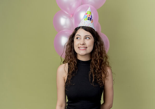 帽子年轻漂亮的女人 卷发 戴着节日帽 手里拿着气球 一边看一边开心地笑着 脸上挂着快乐的表情 站在明亮的墙上庆祝生日派对庆祝气球举行