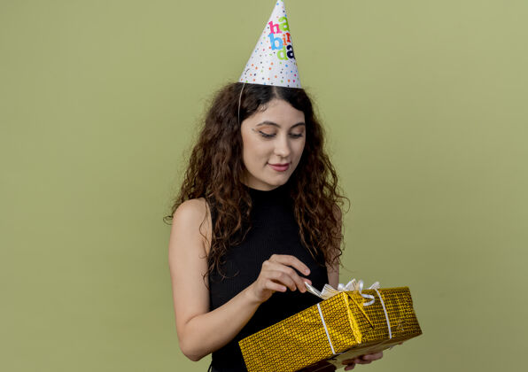 女人一位年轻漂亮的卷发女士戴着节日帽拿着礼盒面带微笑地看着它生日派对理念站在光墙上年轻脸看