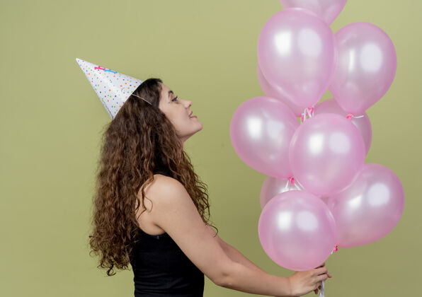 卷曲年轻漂亮的女人 卷发 戴着节日帽 手里拿着气球 快乐而兴奋地站在轻质的墙上空气抱着兴奋