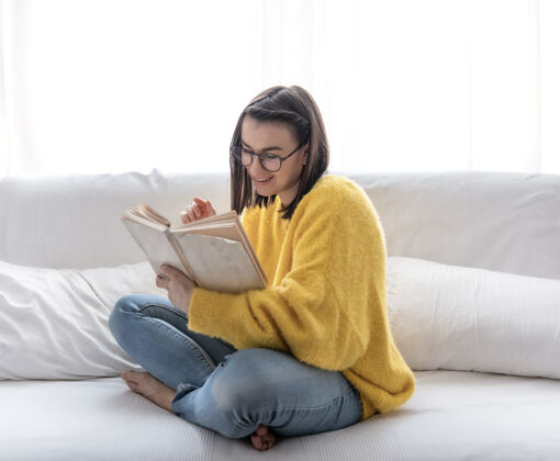 教育穿着黄色毛衣戴着眼镜的时尚黑发女孩在家里的沙发上看书自我发展和放松的概念休闲文学爱好