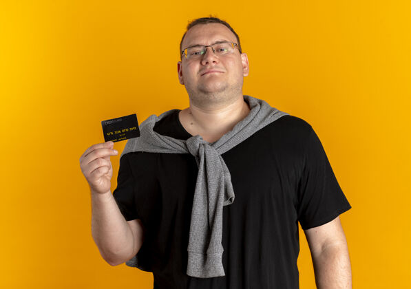 男人戴眼镜的超重男子身穿黑色t恤展示信用卡 面带微笑地看着摄像机站在橙色的墙上站微笑信用
