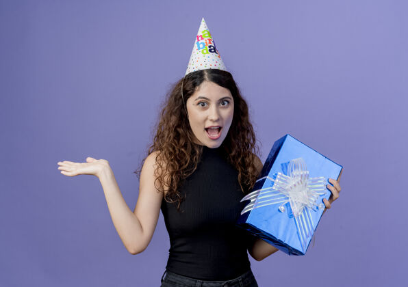 站着一个年轻漂亮的女人 卷发 戴着节日帽 手里拿着生日礼物盒 站在蓝色的墙上 看起来很惊讶 很惊讶生日派对的概念惊奇看着抱着