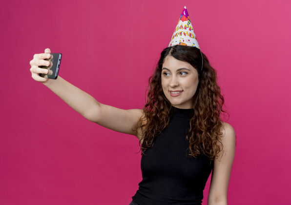 卷发一位年轻漂亮的女士 卷发 戴着节日帽 自拍 微笑着 站在粉色的墙上 欢快地庆祝生日派对欢呼年轻帽子