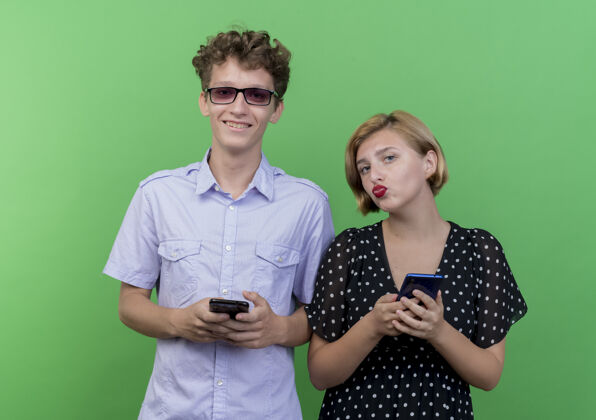 移动年轻漂亮的夫妇手持智能手机微笑着站在绿色的墙上站着男人微笑