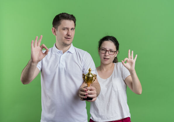 运动年轻的运动情侣站在一起拿着奖杯微笑着表示ok标志站在绿色的墙上年轻情侣站着